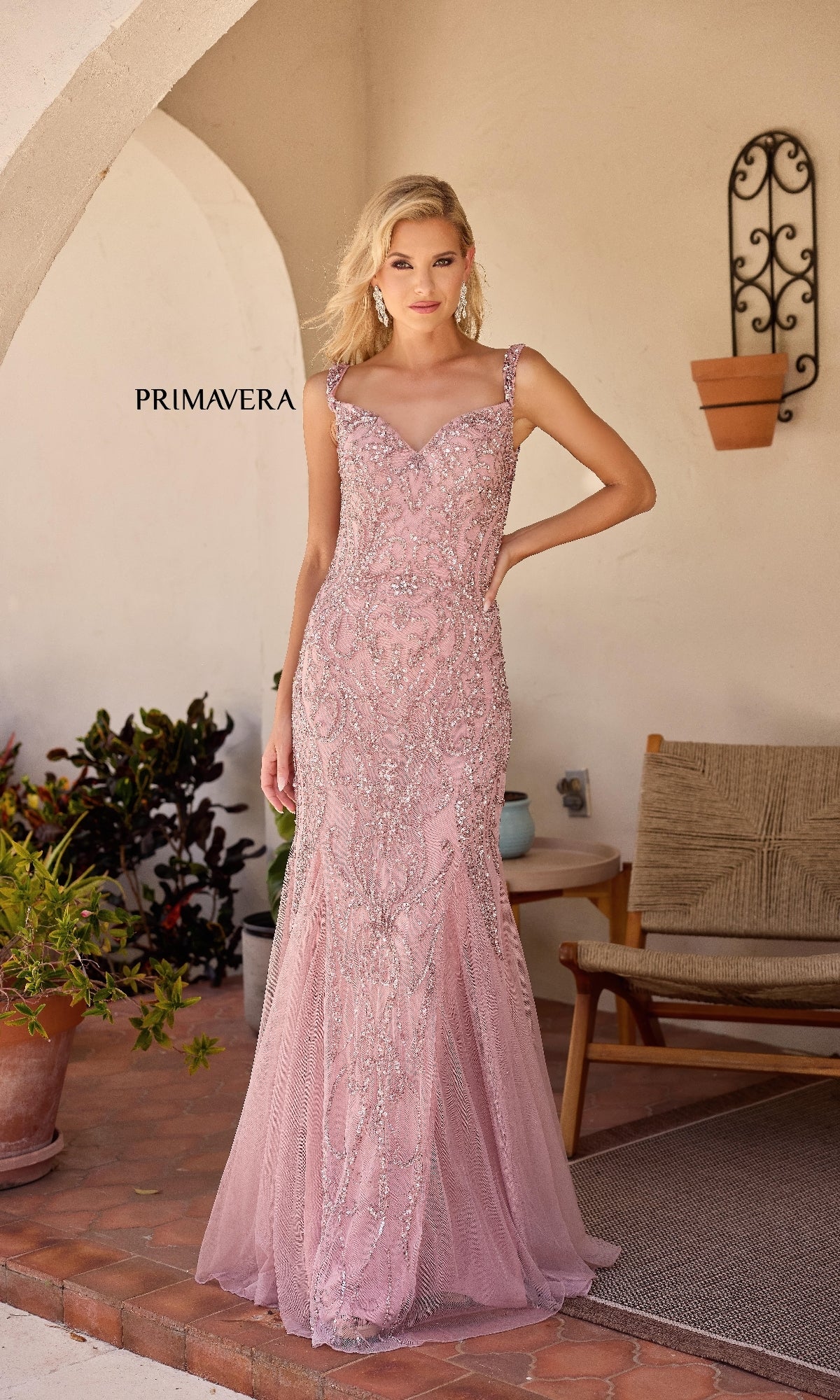 A woman in the Primavera 13143 long formal dress, featuring a light pink hue, sweetheart neckline, and sparkly embellishments, stands on a tiled patio near outdoor furniture and potted plants under an archway.